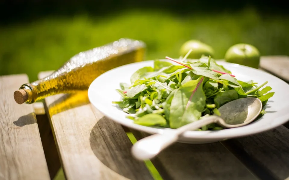 A bowl of green salad with a bottle of olive oil on a wooden table